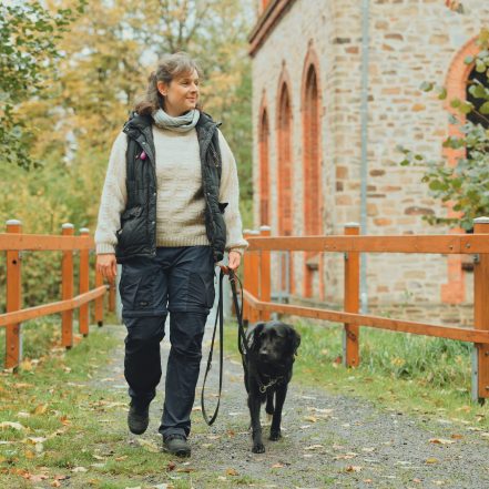 Frau geht entspannt mit einem Hund an der Leine auf ein Weg spazieren. Eine Frau führt einen Hund an der Leine über einen naturbelassenen Weg entlang eines Holzgeländers. Im Hintergrund steht ein Gebäude aus San