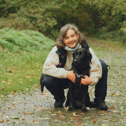 Hundetrainerin Ellie hockt mit ihrem schwarzen Hund auf einem Waldweg. Hundetrainerin Ellie hockt lächelnd auf einem Waldweg und hält ihren schwarzen Hund liebevoll an ihrer Seite.